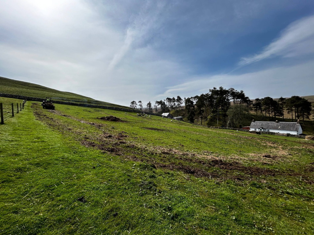 Pastoral scene, likely in a rural area of the British Isles. A small tractor is visible in the foreground, moving along a grassy field that shows signs of recent tilling or land preparation. In the mid-ground, a small, white farmhouse is nestled amongst a stand of dark green pine trees on a hillside. The sky is partly cloudy but bright, suggesting a pleasant day.