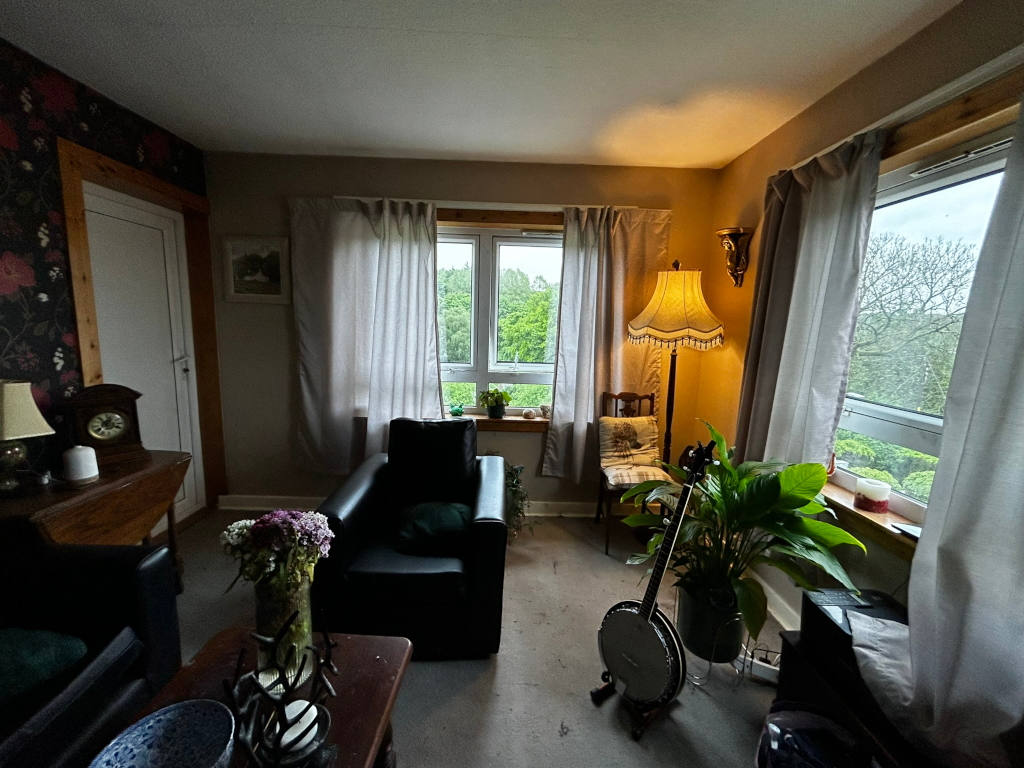 Cosy living room corner. A black leather armchair sits next to a floor lamp with a vintage-style shade.  There's a banjo leaning against a potted plant, adding a musical element. The room has long, light-colored curtains framing windows that show a view of trees, and features dark wood furniture and wallpaper with a floral pattern. The overall mood is calm and slightly rustic, suggestive of a quiet, possibly musical space.