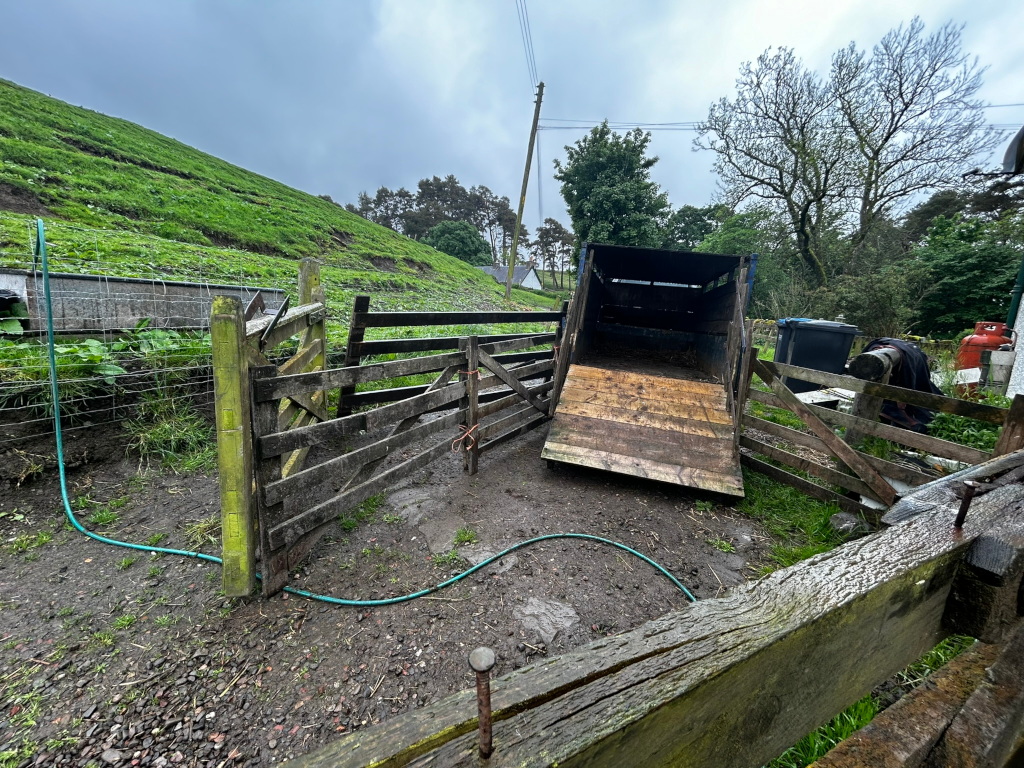 Weathered, wooden livestock trailer, its ramp lowered, resting in a farmyard. The trailer is situated within a rustic, aged wooden enclosure or pen. Green grass and a muddy ground surround the scene. The overall impression is one of rural life and agricultural work. The overcast sky suggests a somewhat dreary day.