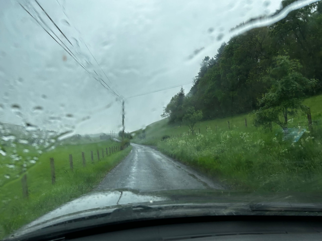 View from inside a car driving down a country road during a heavy rainstorm. Rain droplets cover the windshield, blurring the view somewhat. The road is wet and dark, bordered by lush green fields and trees. The overall atmosphere is moody and somewhat melancholic.