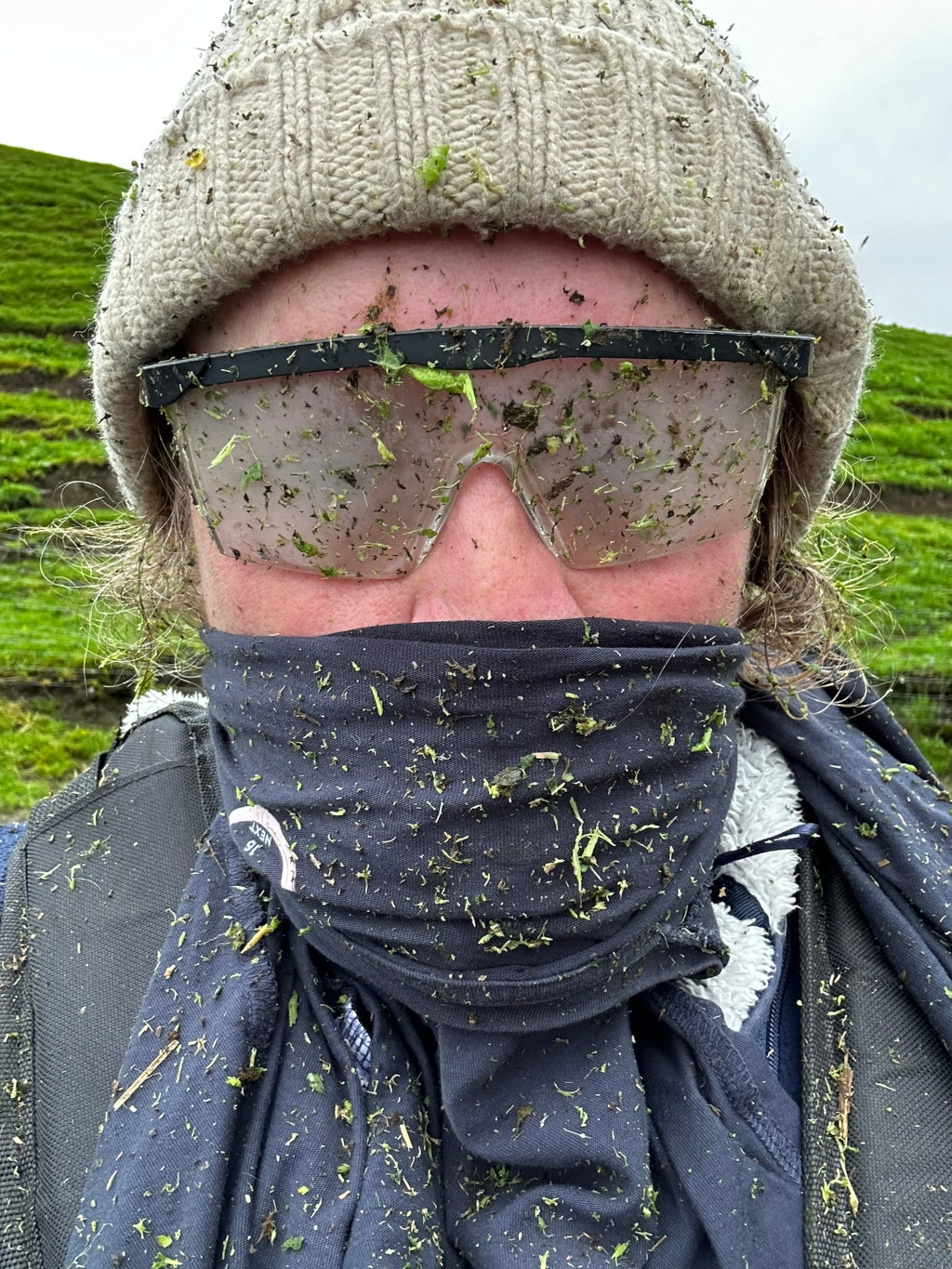 Close-up of a Leonie's face and upper body, heavily covered in grass clippings. Leonie is wearing a beige knit hat, safety glasses, and a dark grey neck gaiter, all liberally dusted with the green debris. The background is a verdant hillside, suggesting Leonie has been involved in landscaping or gardening activities. The overall impression is one of industrious work and a lighthearted acceptance of the messy realities of the job.
