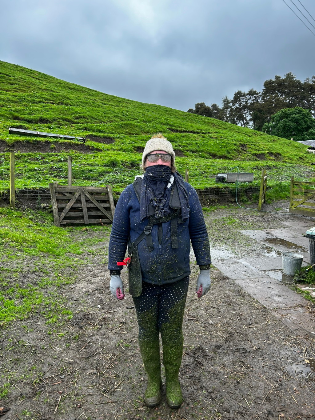 Leonie covered head to toe in what appears to be freshly cut grass, standing in a muddy farmyard. She is wearing protective gear including a face covering, safety glasses, gloves, and tall boots. The background features a lush green hillside and some farm structures. The overall impression is one of a hardworking individual, possibly after completing a task involving lawn care or similar outdoor work. The image has a slightly humorous quality due to Leonie's complete coverage in grass clippings.