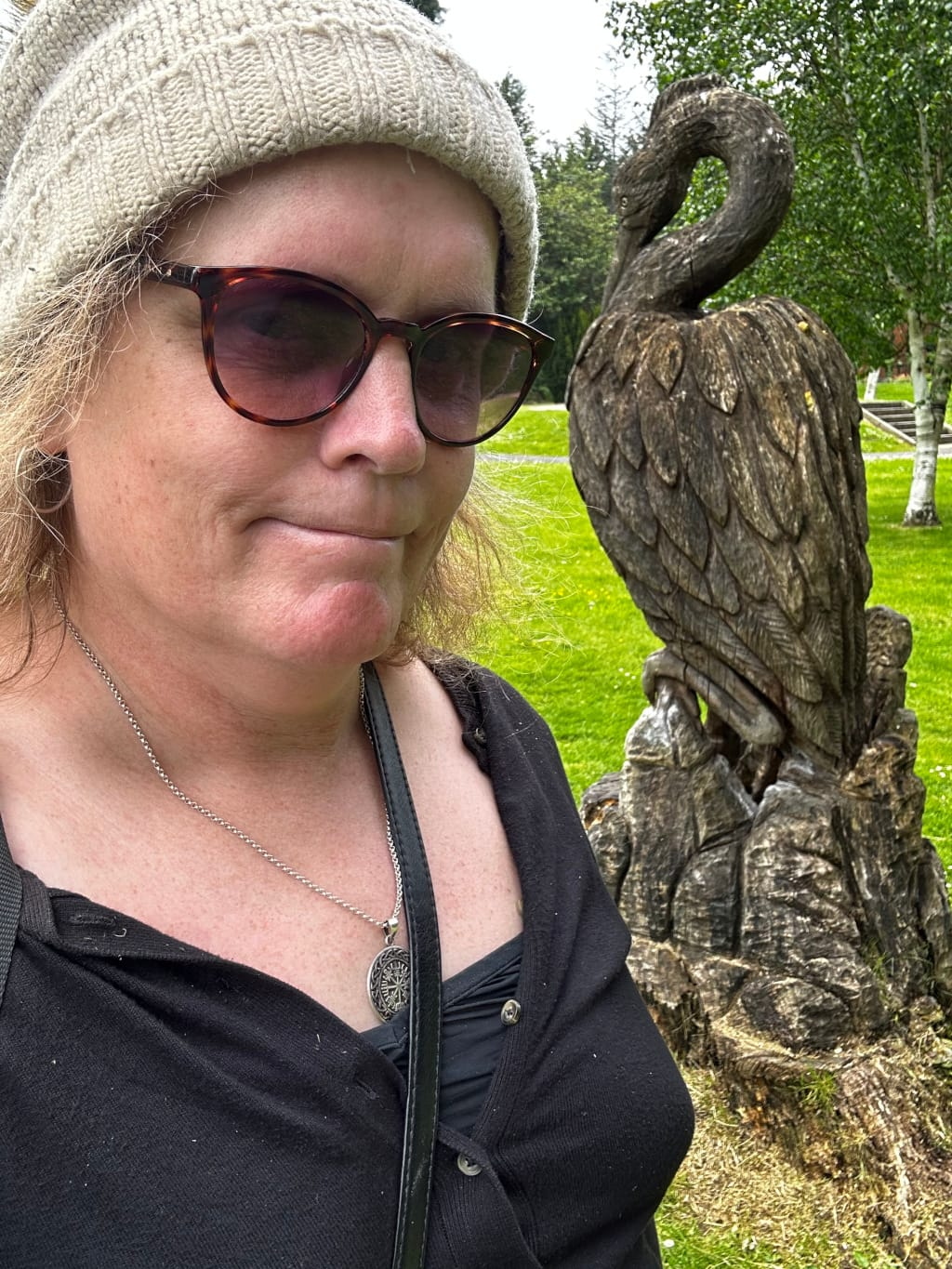 Leonie wearing a beige knit hat and sunglasses, standing in front of a wooden carving of a bird, possibly a heron or crane. Leonie is wearing a black cardigan and a necklace with a circular pendant. The background is a grassy area with some trees.