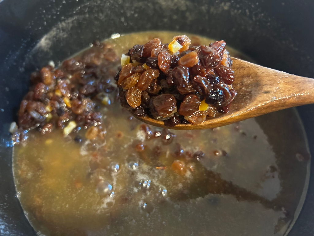 Wooden spoon holding a portion of plump, dark raisins and what looks like candied citrus peel. The spoon is lifting the raisins out of a dark pot containing a simmering liquid and a larger quantity of similar raisins and candied fruit. The liquid appears to be a light brownish-gold colour. The entire scene suggests the preparation of a fruitcake or similar baked good.