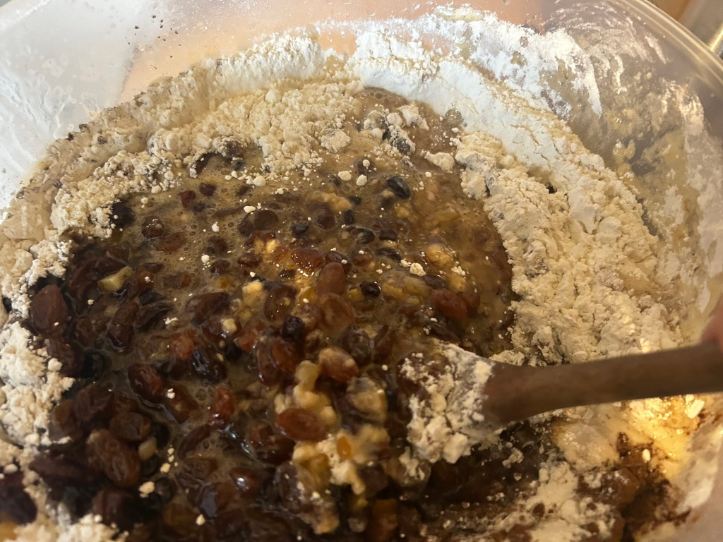 Mixing bowl containing the wet and dry ingredients for a fruitcake. A wooden spoon is partially visible, suggesting the process of mixing is underway. The wet ingredients appear to be a mixture of fruit (raisins, currants, etc.) soaked in liquid, while the dry ingredients consist primarily of flour. The overall appearance is one of preparation for baking.