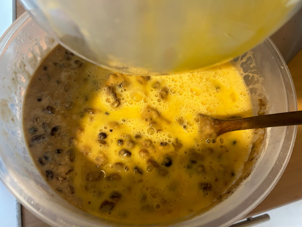 Clear plastic bowl containing a mixture of what appears to be a fruitcake batter. A light-yellow, beaten egg mixture is being poured into the bowl from a separate container, using a wooden spoon to stir. The batter already in the bowl contains dark-coloured fruits and nuts.