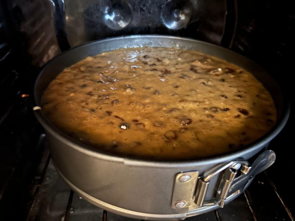 Round cake, possibly a fruitcake, baking in a spring form pan inside an oven. The batter is a light brown colour with visible fruit pieces throughout. The cake appears to be in the middle stages of baking.