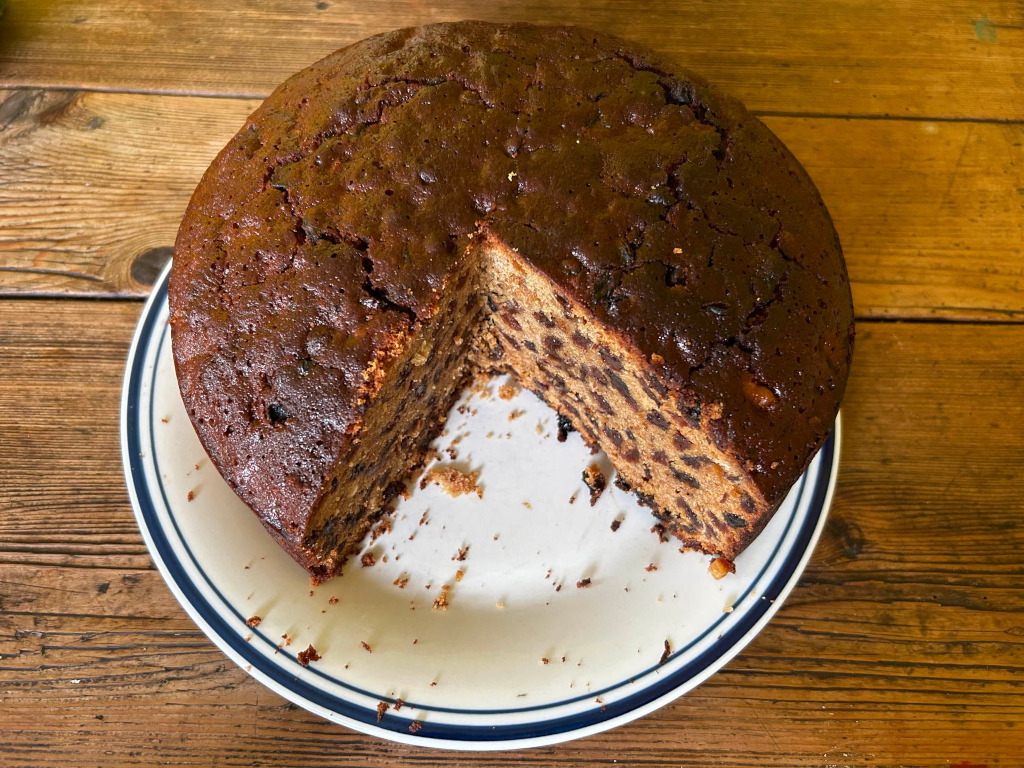 Round fruitcake on a plate. A slice has been cut out, revealing the dense, dark interior studded with dried fruit. The cake has a glossy, dark brown top. The plate is simple, white with a dark blue rim. The background is a rustic wooden table.