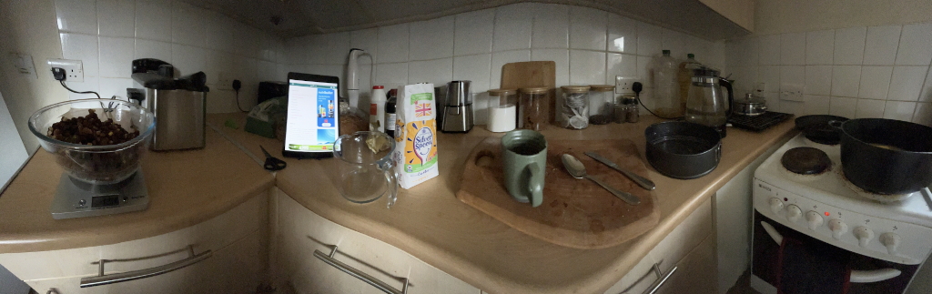 Kitchen counter with various food ingredients and cooking utensils arranged on it. There's a digital tablet displaying a recipe, suggesting baking or cooking preparation is underway. A scale with what appears to be nuts or chocolate chips is present, along with other ingredients like flour, sugar, and tea bags in jars. Several cooking pans and a mug of tea are also visible, and a stovetop with some pans on it is in the background. The overall impression is that of a home kitchen during meal preparation.
