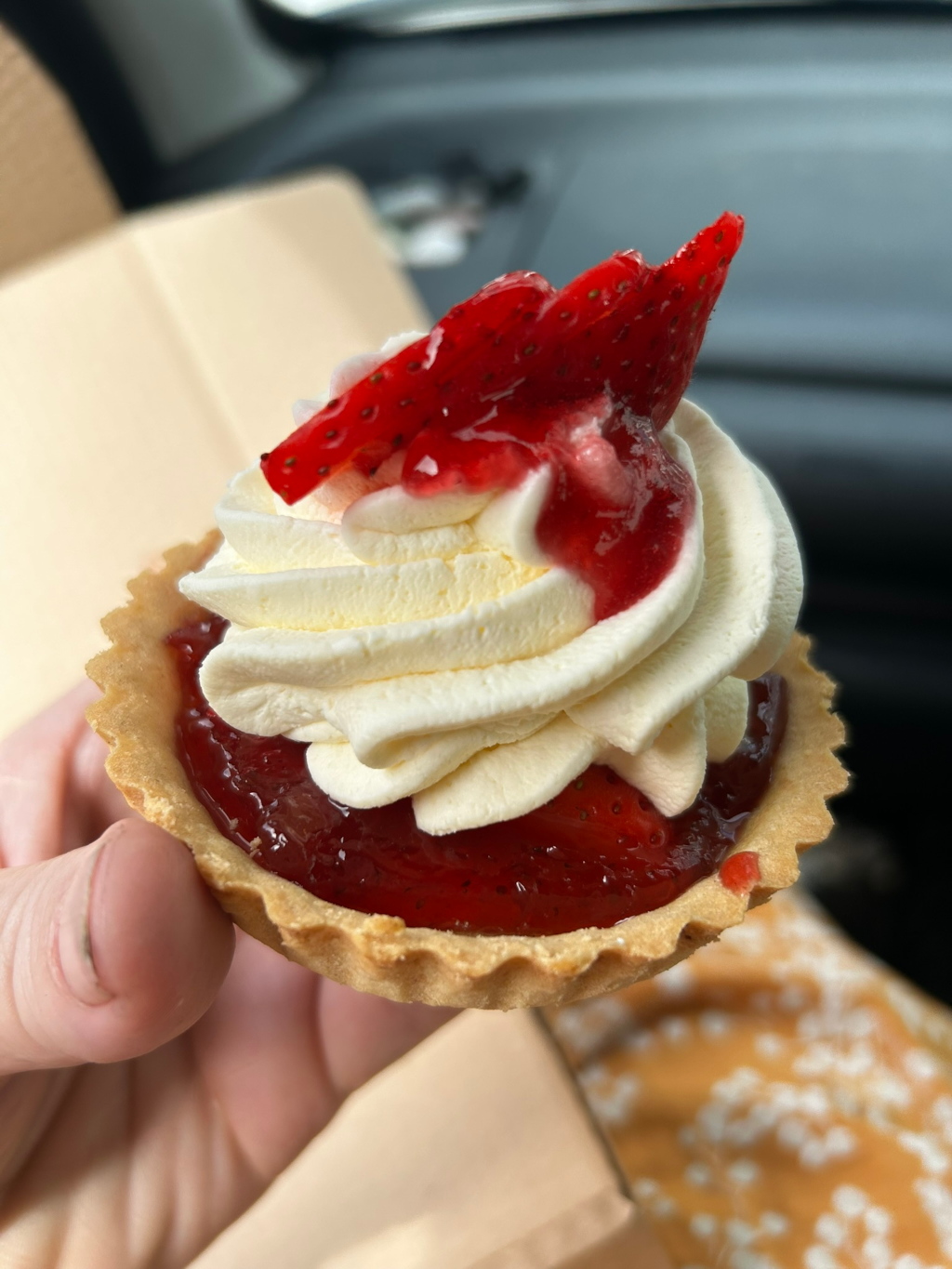 Close-up view of a hand holding a strawberry tart. The tart has a shortbread crust filled with strawberry jam and topped with whipped cream and a fresh strawberry. The background is blurred, showing a car interior and a piece of packaging. The overall impression is one of sweetness and indulgence.
