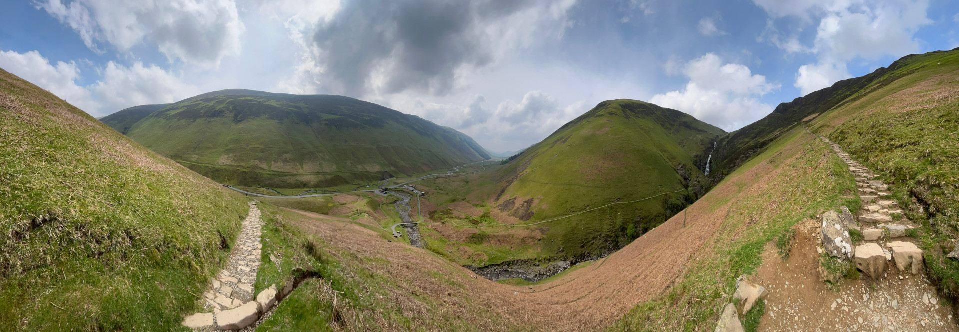 Panoramic view showcases a valley nestled between two verdant hills. A winding road is visible in the valley bottom, alongside a flowing stream. Two stone stairways, one on either side of the image, ascend the hillsides, offering hiking paths up the slopes. A small waterfall is visible on the right-hand hillside, cascading down into the valley. The overall scene is one of tranquil natural beauty, emphasising the rolling hills, the path, and the waterfall.