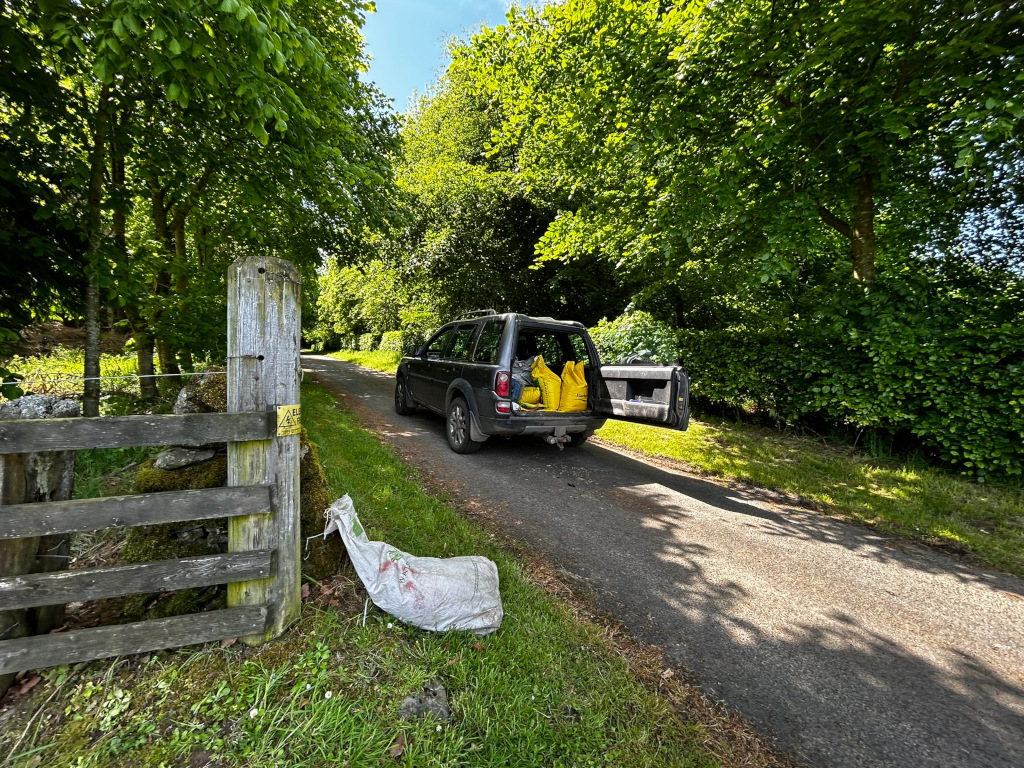 Dark-grey Land Rover parked on a narrow country lane, its rear door open to reveal several yellow sacks inside. The lane is lined with lush green trees and foliage. A rustic wooden fence with moss is visible on the left, and a discarded white sack lies on the grass beside it. The overall scene suggests a rural, possibly agricultural, setting, with the vehicle likely used for transporting goods or supplies.