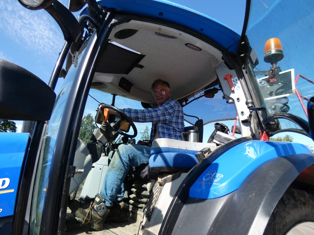 Charlie sitting in the driver's seat of a blue tractor. He is wearing a plaid shirt and jeans. He appears to be middle-aged and has glasses. The tractor is modern and clean. The image is taken from a low angle, looking up at the man and the tractor. The sky is bright blue and sunny. The overall impression is one of a sunny day and a hard-working farmer.