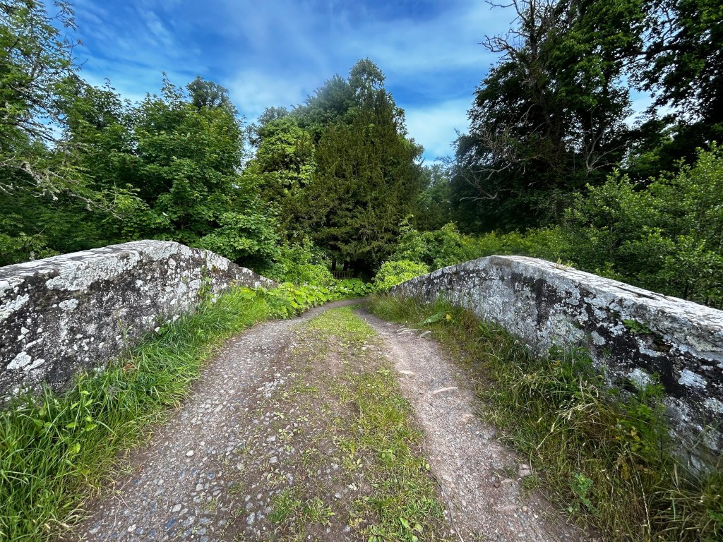 Gravel path leading across a low stone bridge. The bridge is made of weathered grey stone, showing signs of age. Lush green vegetation, including tall grasses and trees, surrounds the bridge and path. The sky is partly cloudy, with blue visible.