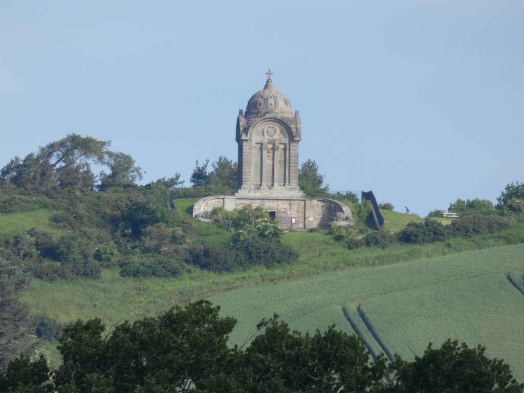 Stone structure, possibly a mausoleum or monument, situated atop a hill. The structure is tall and slender, featuring a domed roof and architectural detailing suggestive of a classical or neoclassical style. It's surrounded by greenery, with lush vegetation covering the hill and surrounding landscape. The lower portion of the image shows a field, possibly a crop field, stretching towards the viewer.