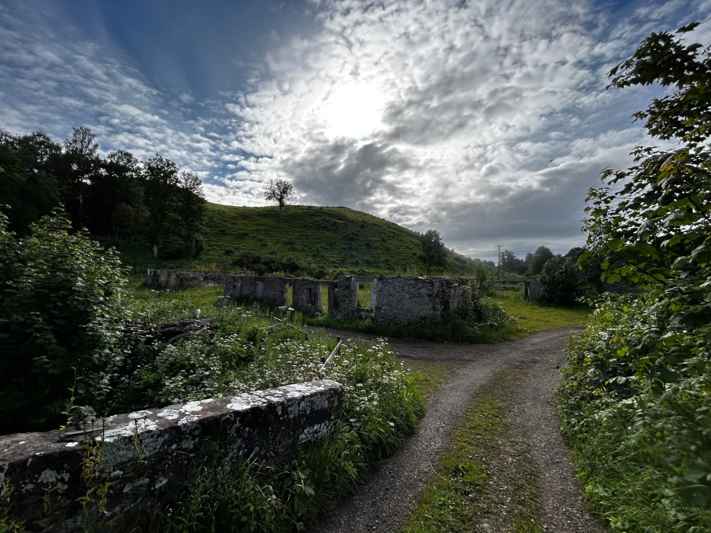 Rural scene featuring a fork in a gravel road leading past the ruins of a stone structure. The ruins appear old and overgrown with vegetation, suggesting abandonment. The background includes a verdant hillside under a partly cloudy sky, with trees lining the landscape.