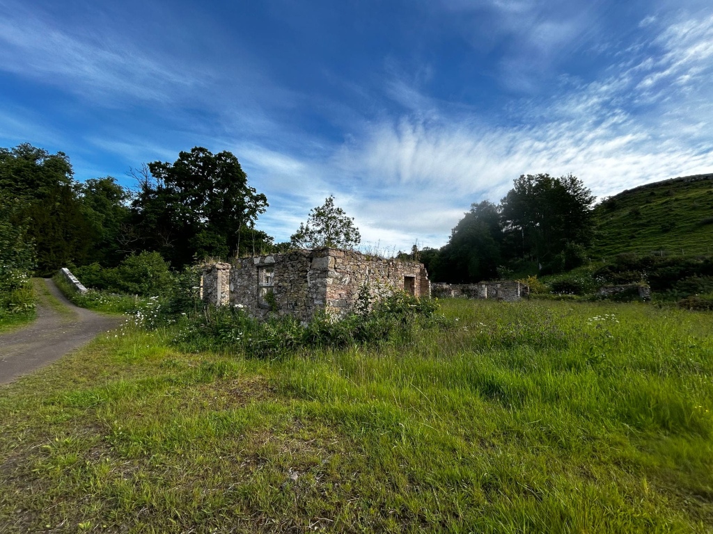 Stone ruin, possibly a small house or outbuilding, partially overgrown with vegetation. It is situated in a grassy field, with a dirt path leading away from it towards a small stone wall and more trees beyond. The background features a verdant hillside under a partly cloudy blue sky. 