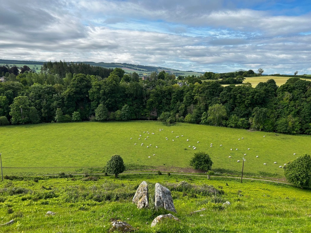 Picturesque landscape view, seemingly from a hilltop, overlooking a verdant pasture dotted with sheep. A line of trees forms a natural border between the pasture and a distant village nestled in a valley, further beyond which rolling hills extend under a partly cloudy sky. In the foreground, two large stones are visible, suggesting a natural or possibly ancient feature.