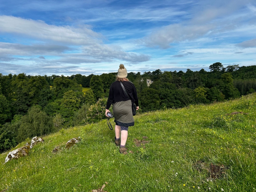 Leonie with her back to the camera, walking away from the viewer across a grassy hillside. She's wearing a dark-coloured top, a green skirt, and a light-coloured beanie. She's carrying a camera. In the background, there's a line of lush green trees and a glimpse of a stone building resembling a castle or manor house, nestled among the trees under a bright, partly cloudy sky.