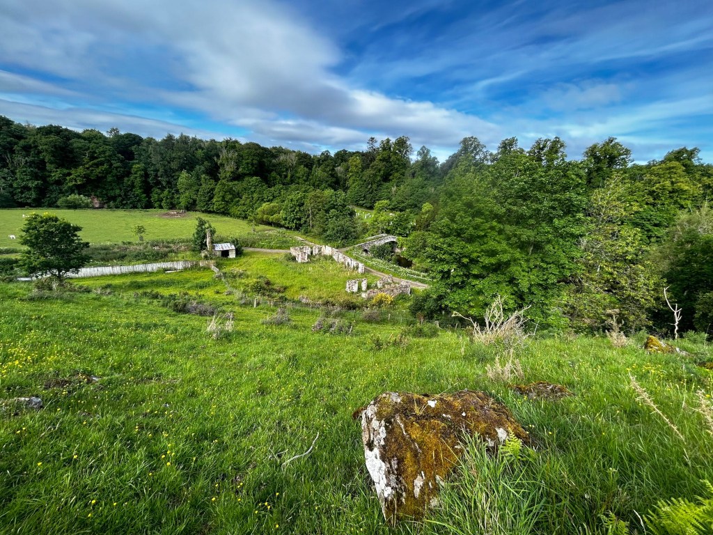 High-angle, long shot of a pastoral landscape. In the foreground is a grassy hillside with a mossy boulder. The mid-ground displays a verdant field and the ruins of what appears to be an old stone structure or wall, possibly part of a former building or enclosure, leading towards a small stone bridge nestled within a lush green wooded area. The background is a dense, verdant forest under a partly cloudy, bright blue sky.