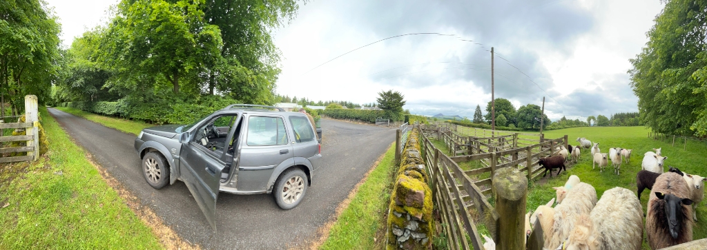 Panoramic view of a rural scene. A silver Land Rover is parked at a crossroads, one road leading to a gate and the other leading further into a grassy field. A flock of sheep is gathered near a wooden fence and stone wall in the field. Lush green vegetation and trees surround the area, and a partly cloudy sky is visible in the background.