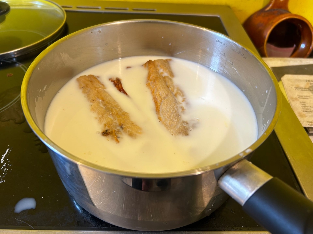 Stainless steel saucepan sitting on a black electric stove top. Inside the saucepan is a creamy white liquid, likely milk or cream, in which two pieces of fried or baked bread are submerged. The bread appears golden brown and slightly crispy. A partially visible lid and a brown ceramic pot are in the background.