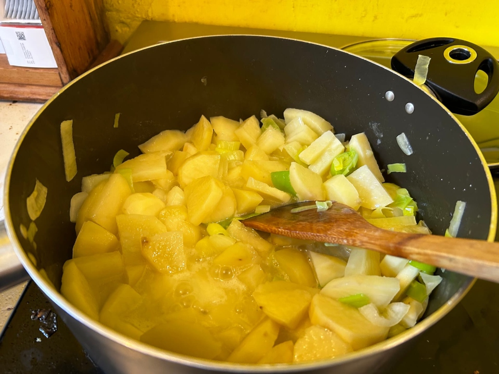 Large, dark-coloured pot on a stove top containing a mixture of cooked potatoes and leeks. A wooden spoon is nestled within the pot of glistening, yellow-hued vegetables. The scene suggests a cooking process underway.