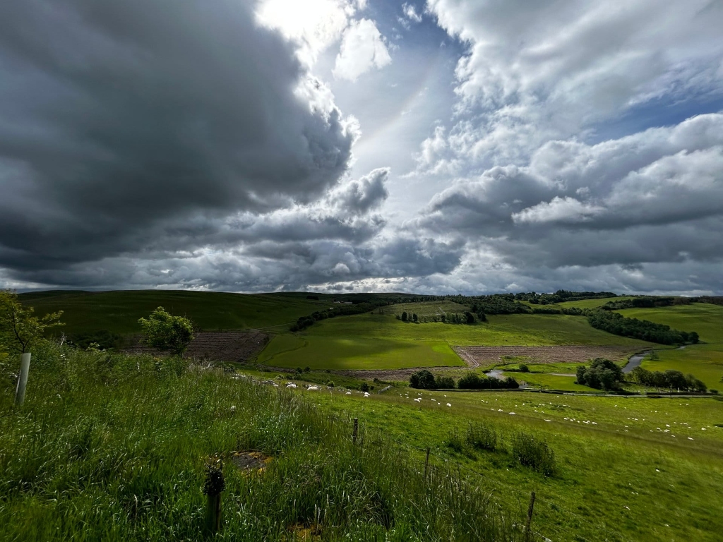 Picturesque landscape under a dramatic sky. Rolling green hills, dotted with sheep and small trees, stretch across the foreground and middle ground. A small stream or river is visible winding through the valley. The sky is filled with dark, heavy clouds, contrasting starkly with the bright sunlight breaking through in patches, creating a moody but beautiful atmosphere. The overall scene evokes a feeling of peaceful rural life juxtaposed with the powerful forces of nature.