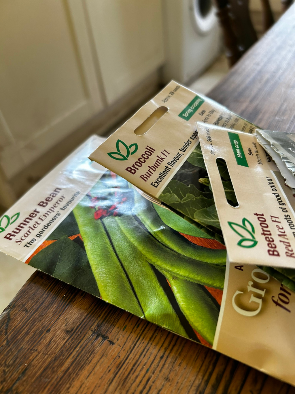 Three seed packets lying on a wooden surface. The packets are for runner beans (Scarlet Emperor), broccoli (Burbank F1), and beetroot (Red Ace F1). The photo appears to be taken in a domestic setting, possibly a kitchen, suggesting preparation for gardening or planting. The focus is on the seed packets themselves, highlighting the varieties and their descriptions. There is no particular narrative or emotional expression beyond the simple depiction of gardening supplies. The moral is implicitly about the cycle of life and the act of nurturing, as is associated with gardening and planting seeds.