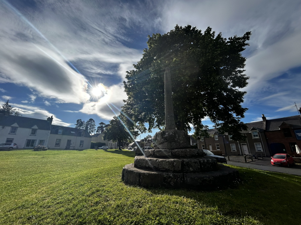 Stone monument, possibly a cross, in a grassy area.  It's situated in front of a row of white and red buildings, which appear to be houses. A large tree partially obscures the monument. The sky is bright and sunny, with streaky clouds. The overall scene suggests a tranquil village or small town setting in the daytime.