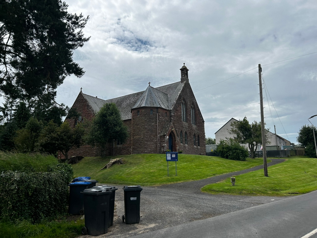 Stone church situated on a grassy hill. The church is a reddish-brown stone with a steeply pitched roof and a small spire. To one side, there are several black garbage bins. In the background, there are some modern houses and lush green vegetation. 