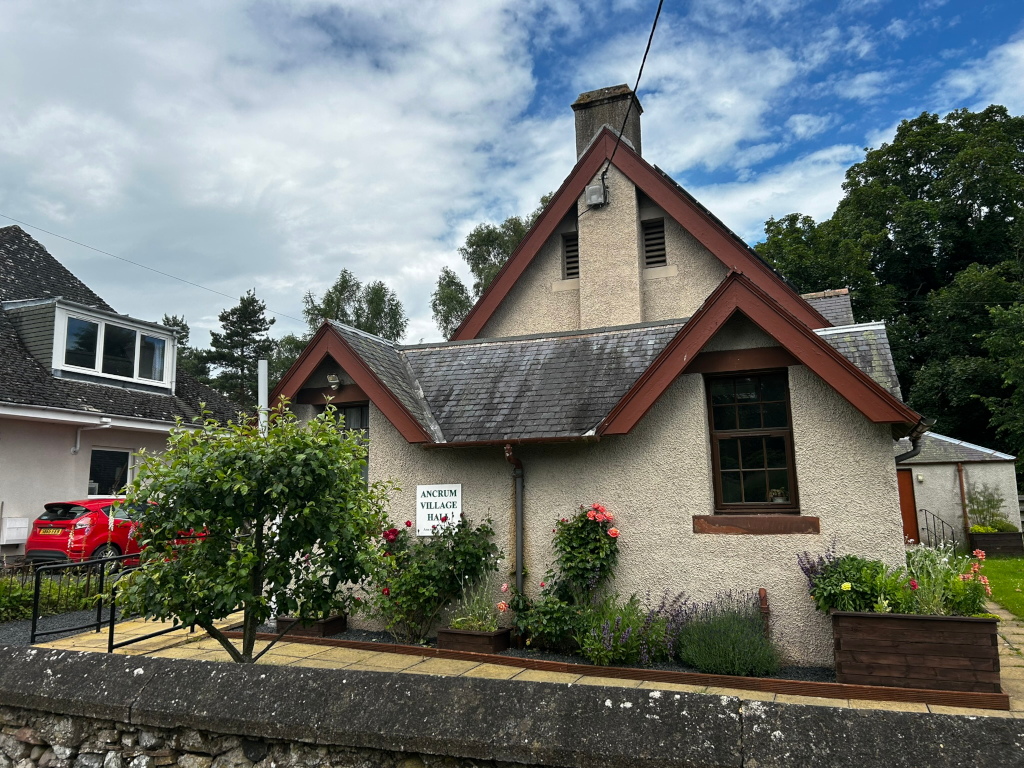 Ancrum Village Hall, a small, single-story building with a steeply pitched roof and reddish-brown trim. The building is light beige with dark windows.  It's surrounded by well-maintained landscaping, including shrubs, flowers in planters, and a small tree in front. A red car is parked in front of an adjacent building. The sky is partly cloudy.