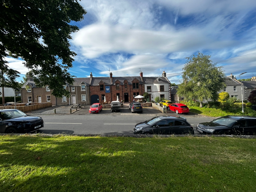 Row of terraced houses on a sunny day. Several cars are parked in front of the houses, including a red car, a yellow car, and several dark-coloured cars. A small, grassy area is in front of the houses, and a larger grassy area is in the foreground of the image, suggesting a slightly elevated viewpoint. The sky is mostly blue with fluffy white clouds.