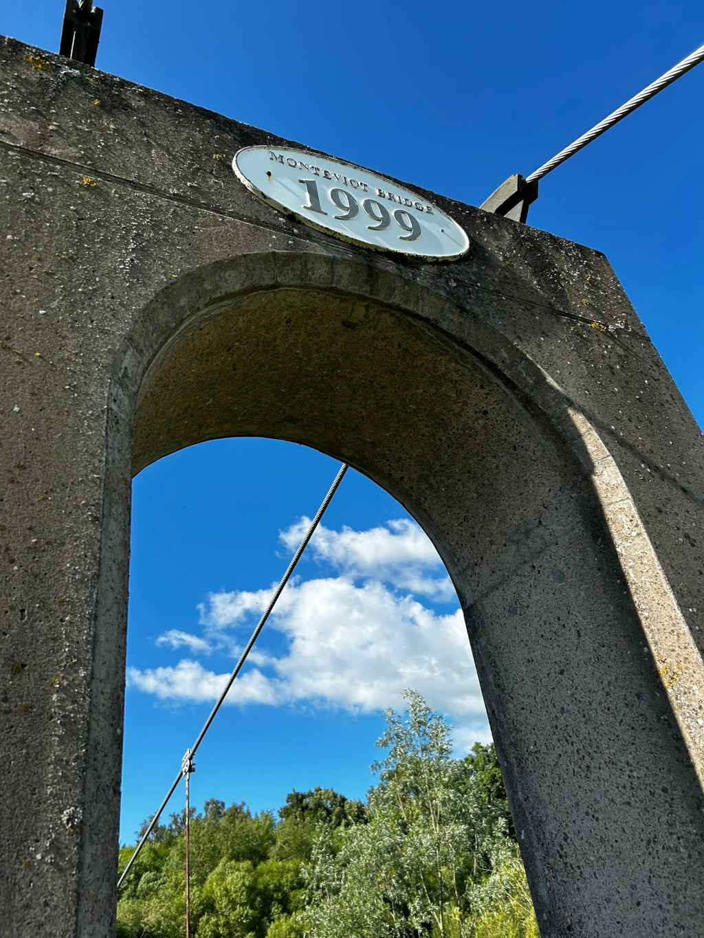 Close-up view of a concrete pillar of the Monteviot Bridge, built in 1999. The pillar features an arched opening that frames a clear blue sky with fluffy white clouds. A suspension bridge cable stretches diagonally across the frame, partially obscured by the arch. Lush green trees are visible in the distance, below the arch.