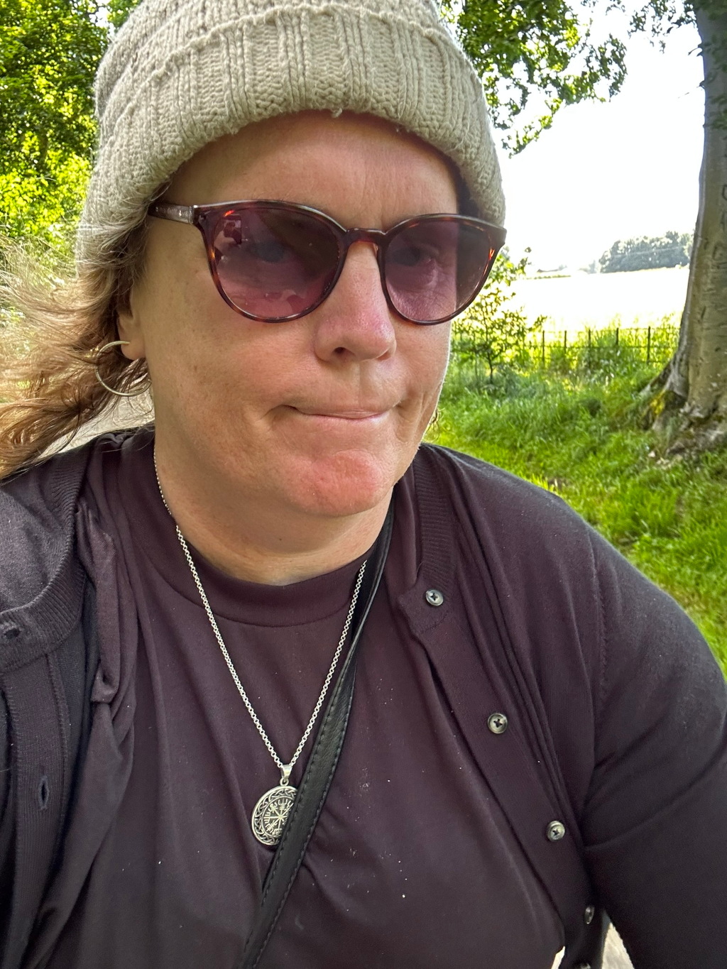 Close-up selfie of Leonie outdoors. She's wearing a beige knit cap, brown sunglasses, a dark-coloured henley shirt, and a necklace with a circular pendant. Her expression is somewhat serious or neutral. The background is slightly blurred but shows lush green vegetation and a glimpse of a field or open area in the distance.
