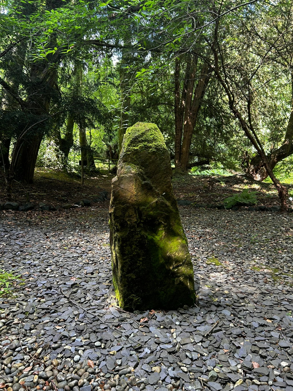 Moss-covered standing stone situated in a clearing within a lush, green woodland. The ground around the stone is covered with dark grey slate chips and pebbles. The overall effect is one of serene tranquillity and possibly hints at a contemplative or spiritual setting.