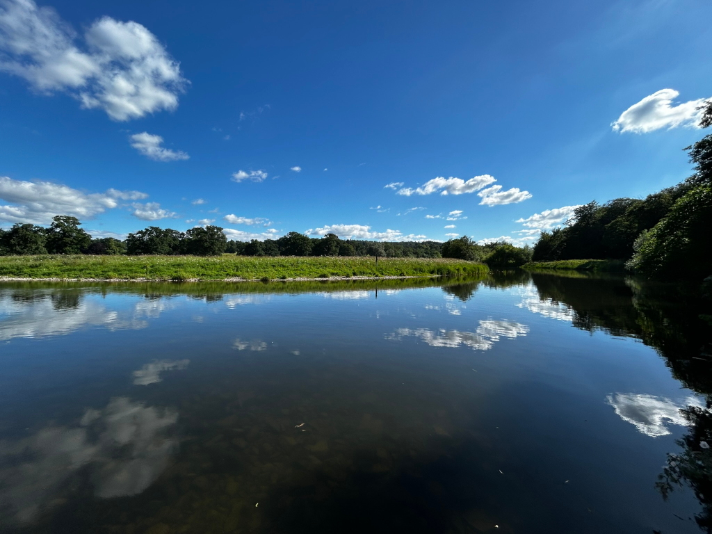 Tranquil river scene under a bright, sunny sky. The river is calm, reflecting the fluffy white clouds and blue sky. Lush green vegetation lines the riverbanks, with trees visible in the distance.