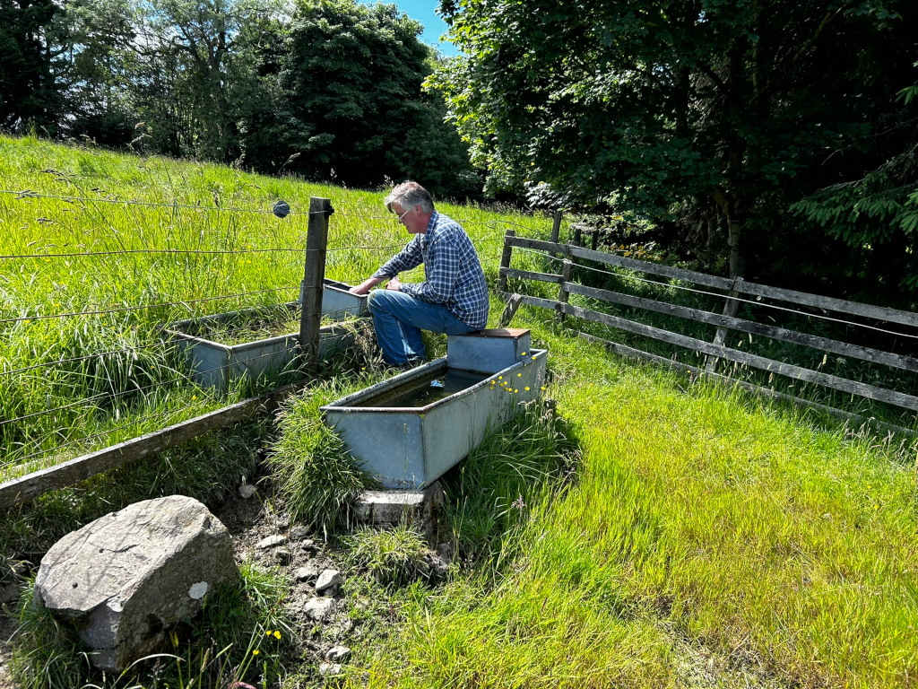 Charlie in a plaid shirt sitting next to two galvanised steel troughs, possibly animal watering troughs, in a grassy field. He is leaning over one of the troughs, possibly checking the water level or cleaning it. A wooden fence runs along the edge of the field. A large stone is visible in the foreground.