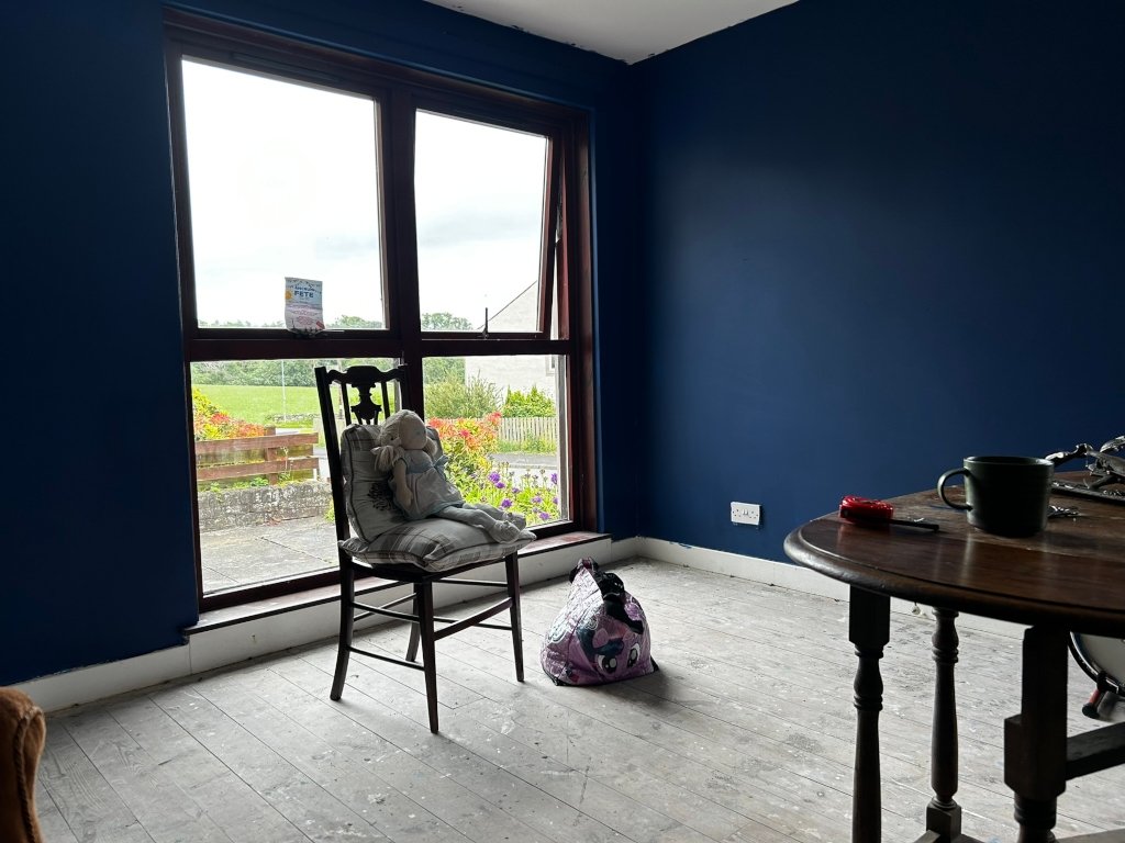 Room with dark blue walls and a large window overlooking a garden. A wooden chair sits near the window with a doll seated on it. A small bag lies on the floor beside the chair. A dark wooden table with various items, including a mug, is partially visible in the foreground. The floor appears to be unfinished wood. The overall impression is one of a room undergoing renovation or decoration.