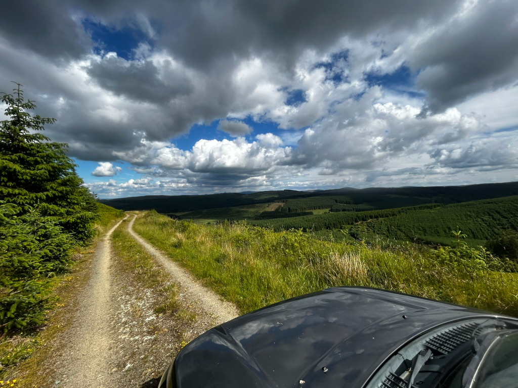 Gravel road winding through a lush green landscape, leading towards a distant valley under a partly cloudy sky.  A dark-coloured car's hood is partially visible in the bottom right corner, suggesting a journey or road trip. The overall feeling is one of serene countryside scenery.