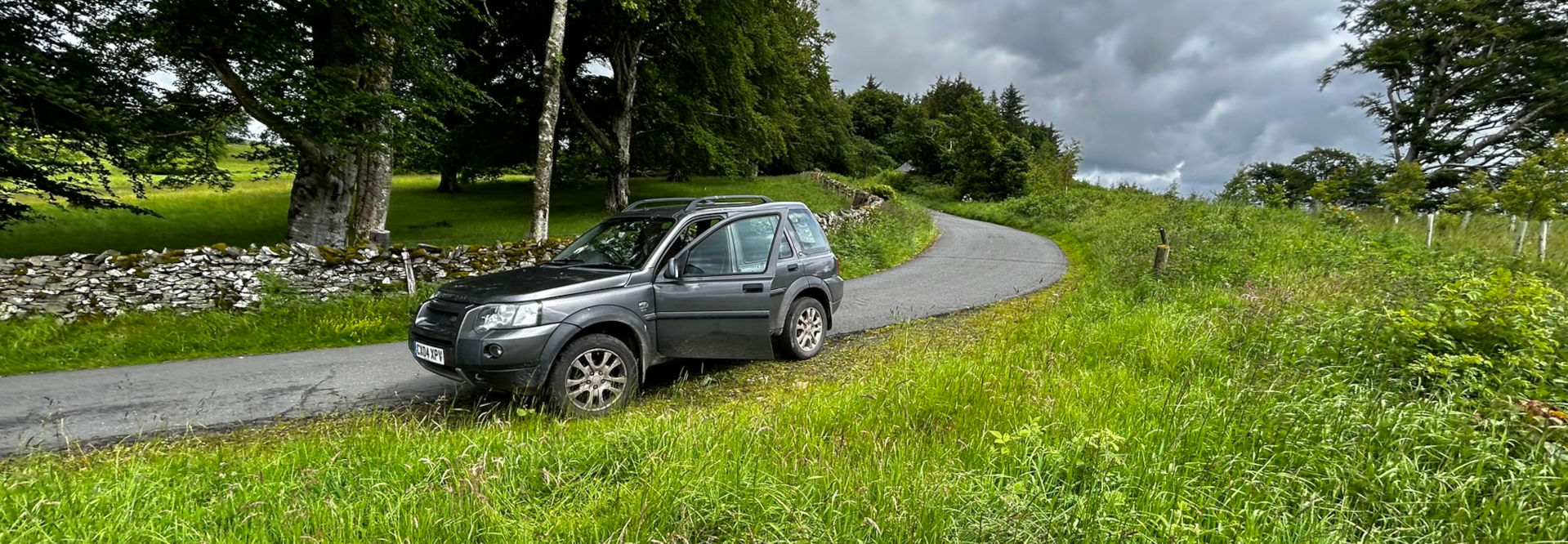 Dark grey Land Rover Freelander parked on the side of a country road. The road curves gently to the right, disappearing into the distance towards a line of trees on a hill. The surrounding landscape is lush green, with tall grass and a low stone wall running alongside the road.  The sky is overcast, with dark stormy-looking clouds.