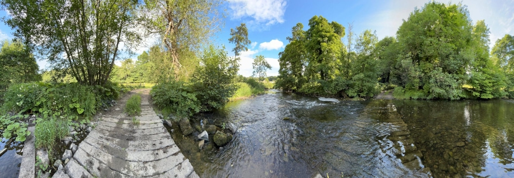 Panoramic view of a tranquil river scene. A stone path or ford crosses a shallow section of a clear river, bordered by lush green vegetation and mature trees. The sky is partly sunny, suggesting a pleasant day. The overall impression is one of peace and serenity in a natural setting.