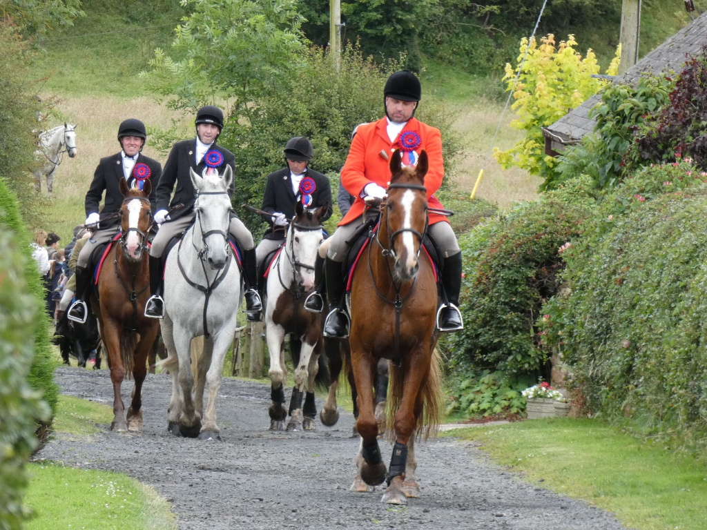 Four people on horseback riding down a country lane. They are dressed in formal riding attire, with the lead rider wearing a bright red coat. All riders appear to be wearing rosettes or ribbons on their jackets, suggesting they may have participated in some sort of equestrian competition or event. The setting is rural, with hedges and greenery lining the road. There are also other people visible in the background, and another horse can be seen further up the lane. The overall atmosphere is one of quiet formality and possibly a celebratory procession.