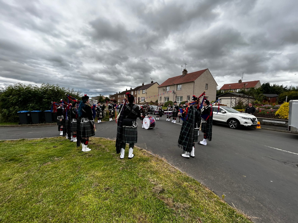 Bagpipe band, predominantly dressed in kilts, performing in a residential street. They are positioned in a line, facing away from the camera towards a small group of drummers. The setting appears to be a small town or village, with modest houses lining the street. The sky is overcast and cloudy.