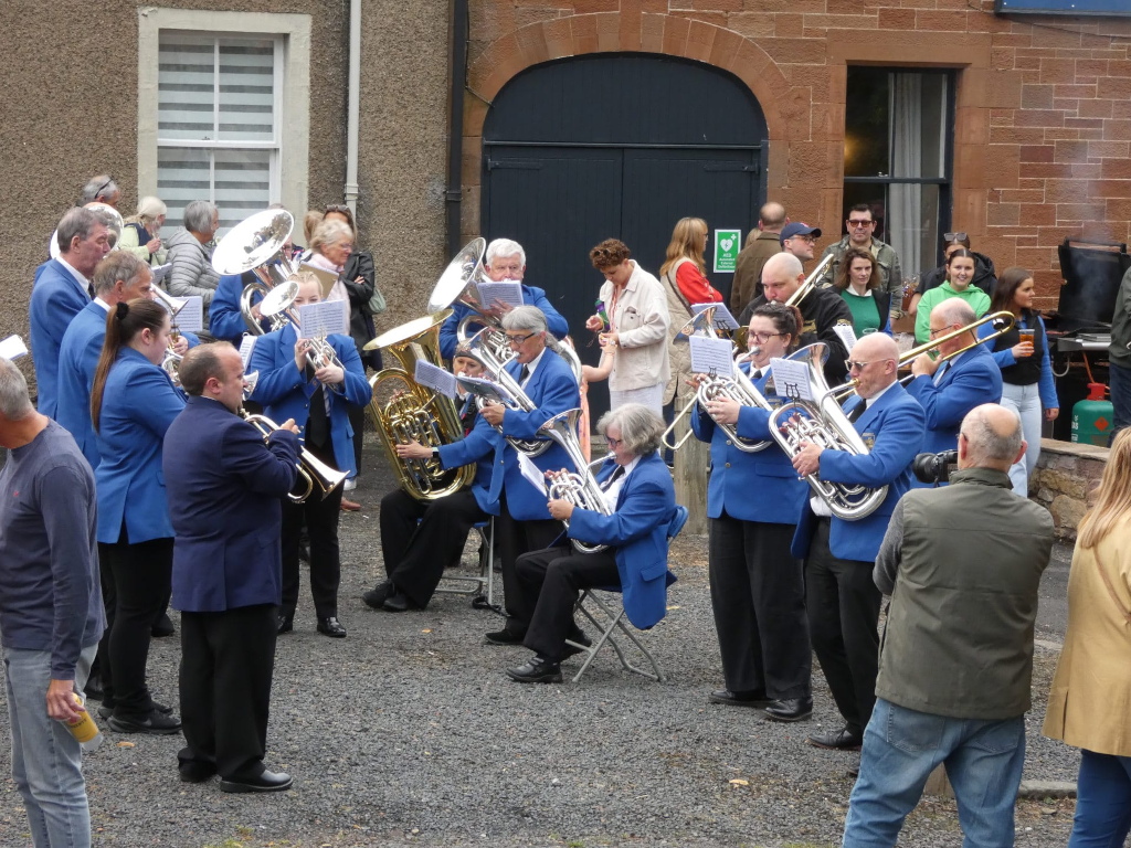 Brass band, predominantly dressed in blue uniforms, performing outdoors. They are positioned in front of a stone building, with a mix of onlookers and other individuals standing in the background. The band members are playing various brass instruments including euphoniums, trumpets, cornets, and a tuba.