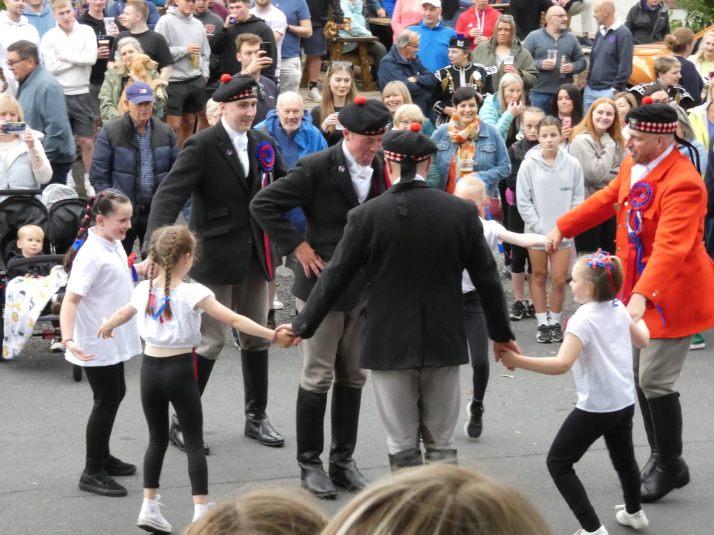 Group of people, predominantly dressed in traditional Scottish attire, participating in what appears to be a cultural event. Several young girls in white shirts and black leggings are being led in a dance or procession by men wearing kilts and jackets. A man in a bright orange jacket seems to be officiating or leading the event. There's a large crowd of onlookers in the background. The overall atmosphere suggests a festive and communal celebration of Scottish culture.