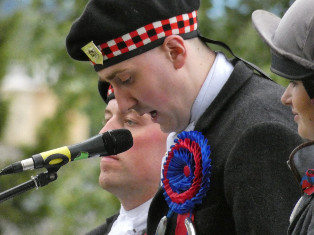 Close-up of a man wearing a black beret with a red and white chequered band, a grey jacket, and a blue and red rosette. He is speaking into a microphone. Behind him and partially visible are parts of two other people. One appears to be wearing a similar style of hat, implying a possible collective event, possibly a competition or ceremony. The overall feel is one of formality and possibly a public speaking event, perhaps related to a traditional Scottish cultural activity given the man's hat.