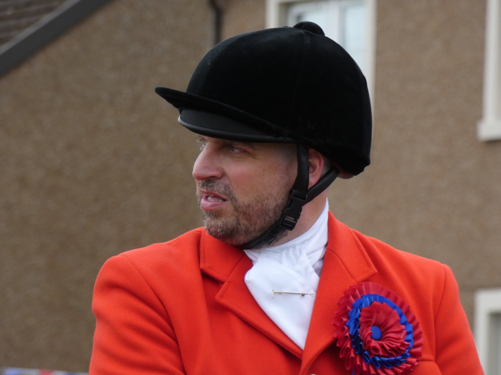 Close-up of a man wearing a black riding helmet and a bright red hunting coat. He has a white shirt and cravat visible under the coat. A blue and red rosette is pinned to his lapel. The man appears to be outdoors, with a neutral-coloured building visible in the background. His expression is serious, almost pensive, and he's looking off to his side. The overall impression is one of formality and perhaps participation in a hunt or equestrian event.