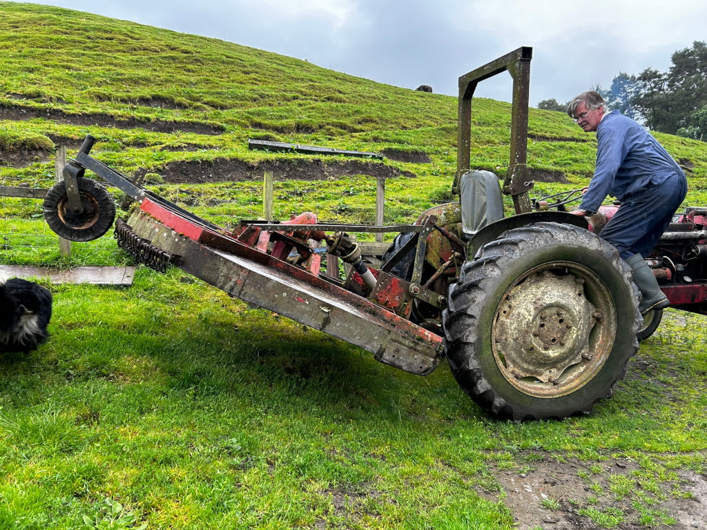 Charlie operating a vintage, red and grey tractor with a side-mounted rotary mower. The tractor is partially tilted, with the mower extended on the ground.  A black and white border collie rests in the grass nearby, seemingly observing the work.
