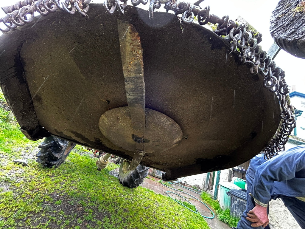 Large, circular, rusty, and dirty metal object, possibly a piece of agricultural machinery like a flail mower or rotary cutter. It's lifted up, revealing its worn condition and a chain mechanism attached to its perimeter. The scene is outdoors, on grass, with a person partially visible in the bottom right corner suggesting maintenance or repair work. Rain or water droplets are visible on the object.